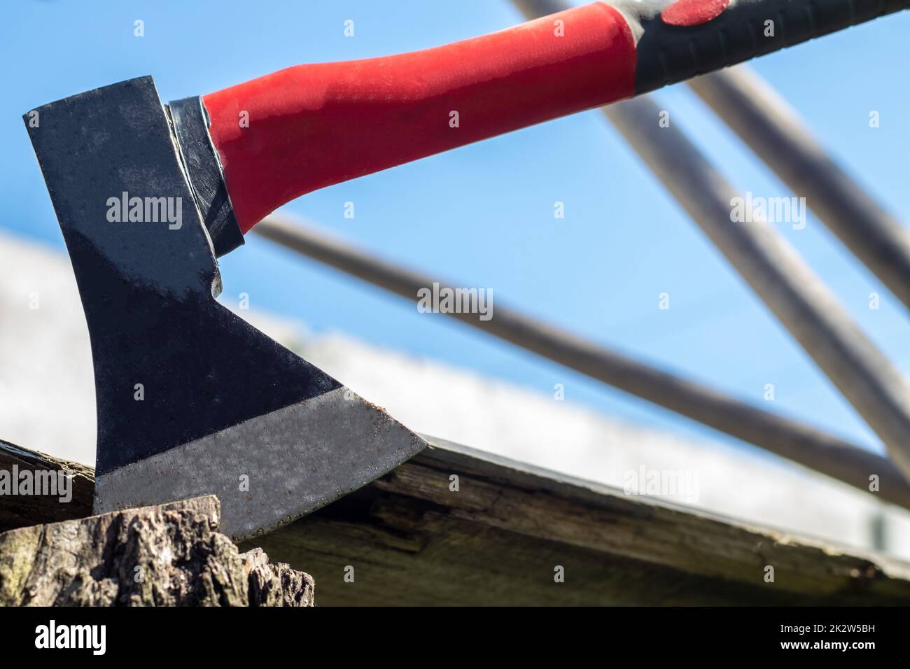 A new modern ax with a red handle sticks out of a wooden stump against a green meadow on a summer sunny day. The ax stuck into the stump. Ax blade in a log. Stock Photo