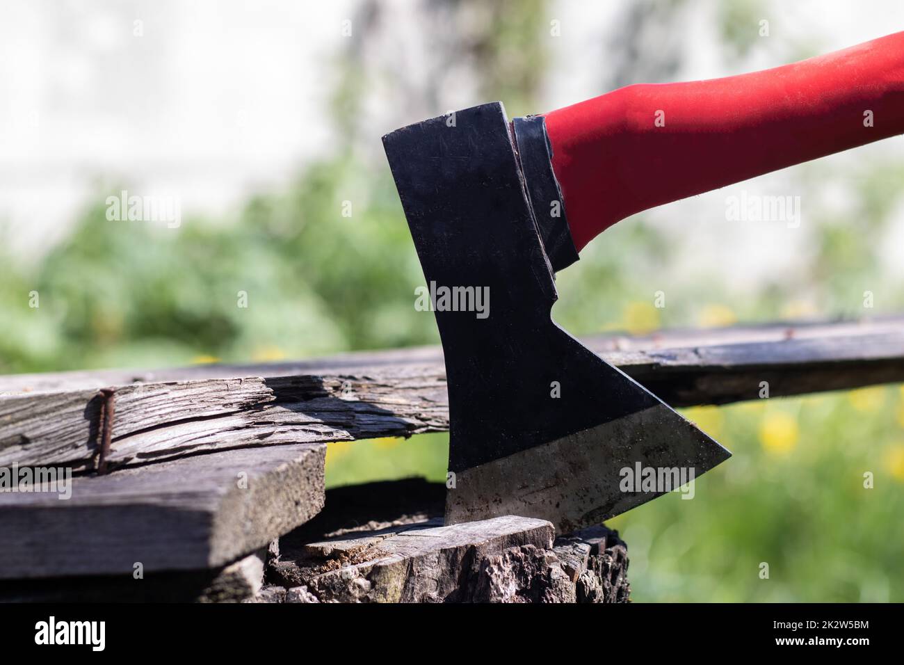 A new modern ax with a red handle sticks out of a wooden stump against a green meadow on a summer sunny day. The ax stuck into the stump. Ax blade in a log. Stock Photo