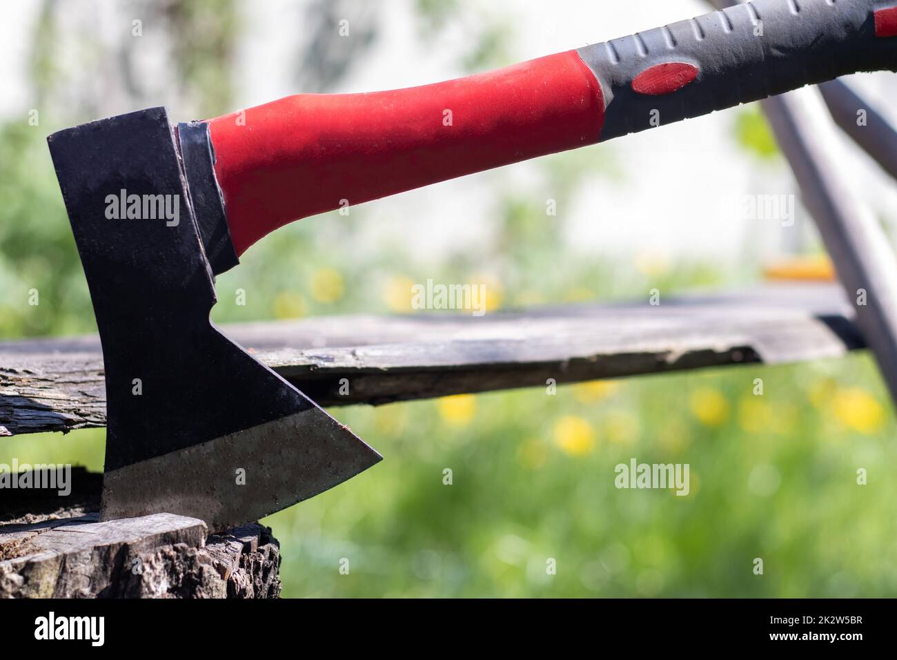 A new modern ax with a red handle sticks out of a wooden stump against a green meadow on a summer sunny day. The ax stuck into the stump. Ax blade in a log. Stock Photo