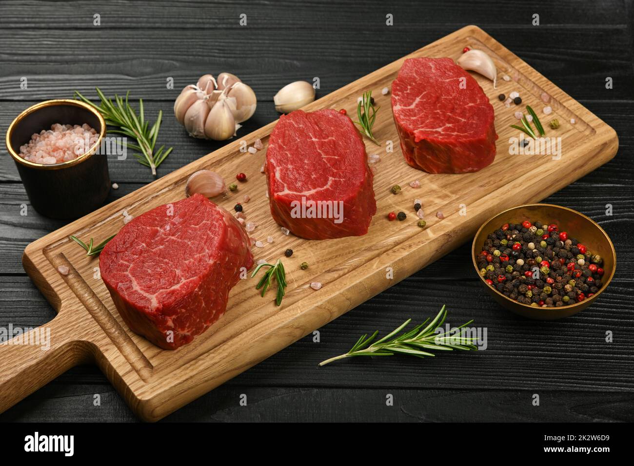 Close up three aged prime marbled raw tenderloin or fillet mignon beef steaks on brown oak wood cutting board, with spices, over black wooden table ba Stock Photo