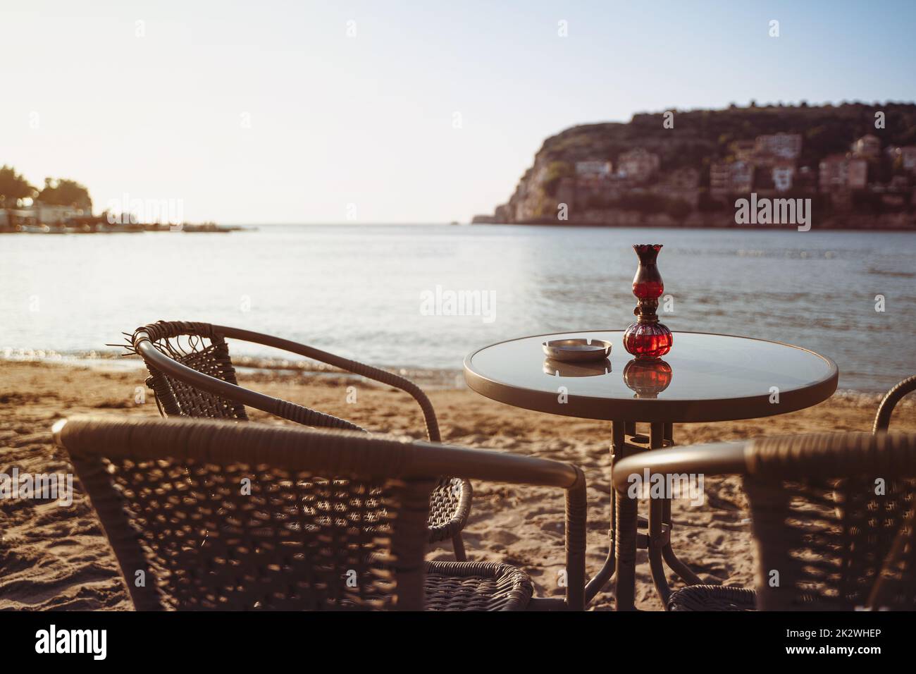 rear view closeup of empty cafe table and chairs on sandy beach with ashtray and candle light in front of sea golf with clear sky before sunset Stock Photo