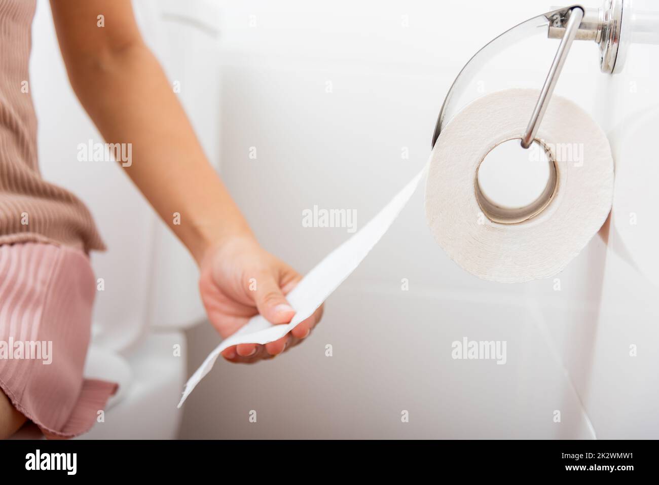 Closeup hand pulling toilet paper roll in holder for wipe.Closeup hand pulling toilet paper roll in holder for wipe, woman sitting on toilet she taking and tearing white tissue on wall to towel clean in bathroom, Healthcare concept Stock Photo