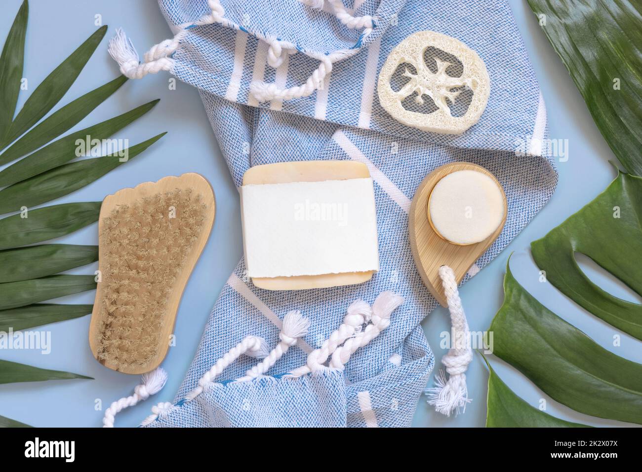 Handmade soap bar on blue bath towel near tropical leaves top view