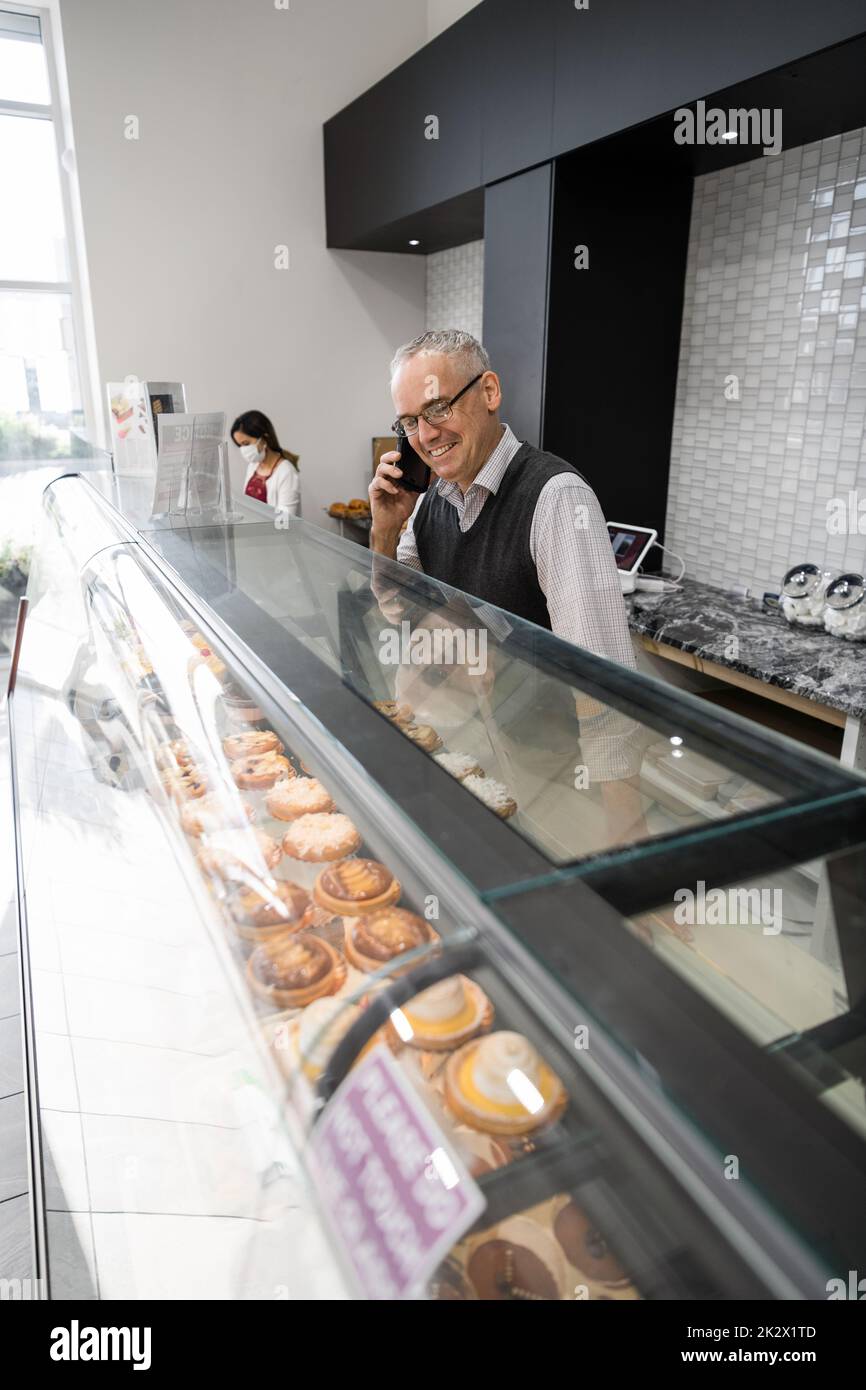 Happy male bakery owner talking on smart phone behind display case Stock Photo