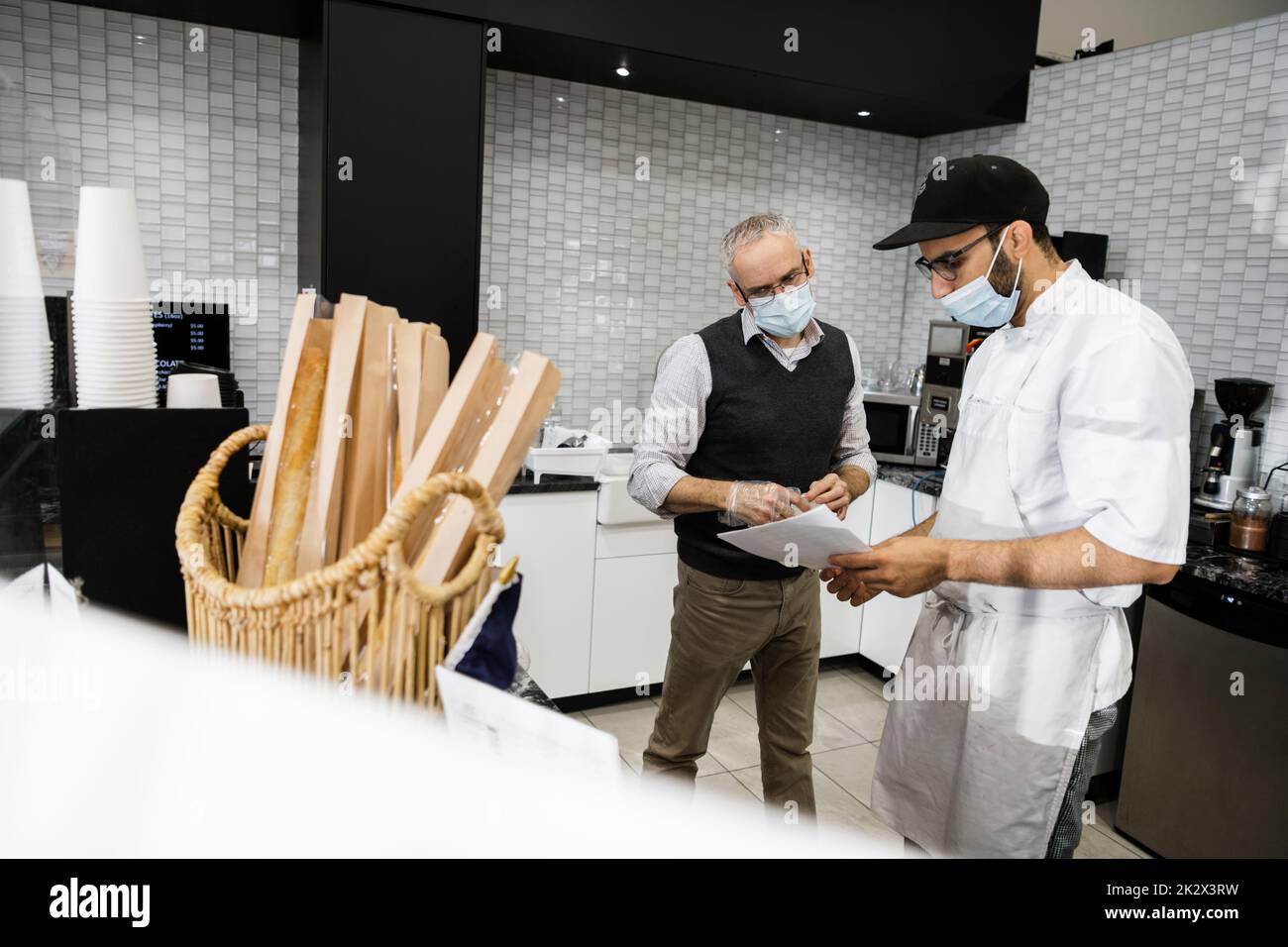 Male baker and bakery owner in face masks reviewing order paperwork Stock Photo