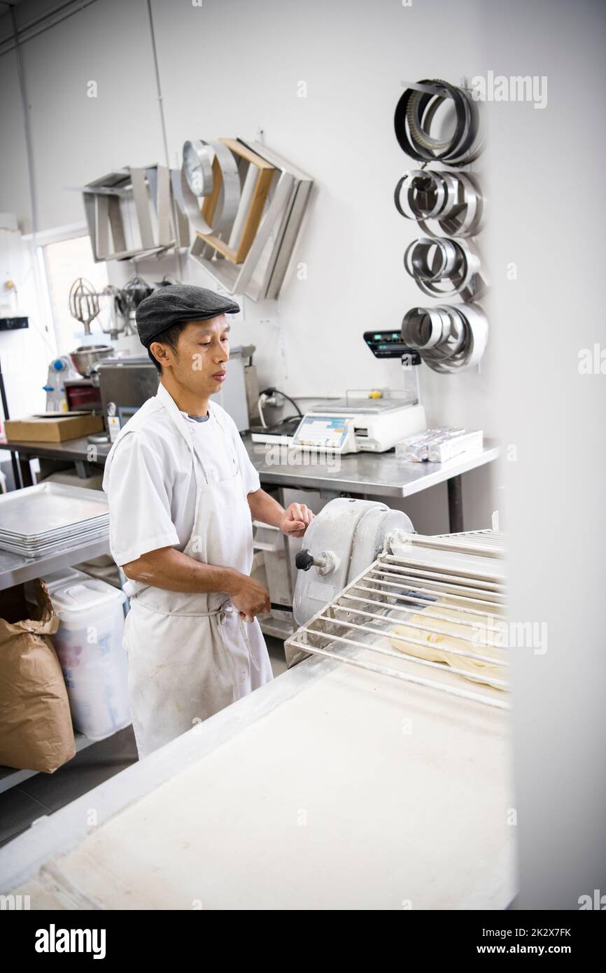 Male baker using commercial equipment to roll dough in bakery kitchen