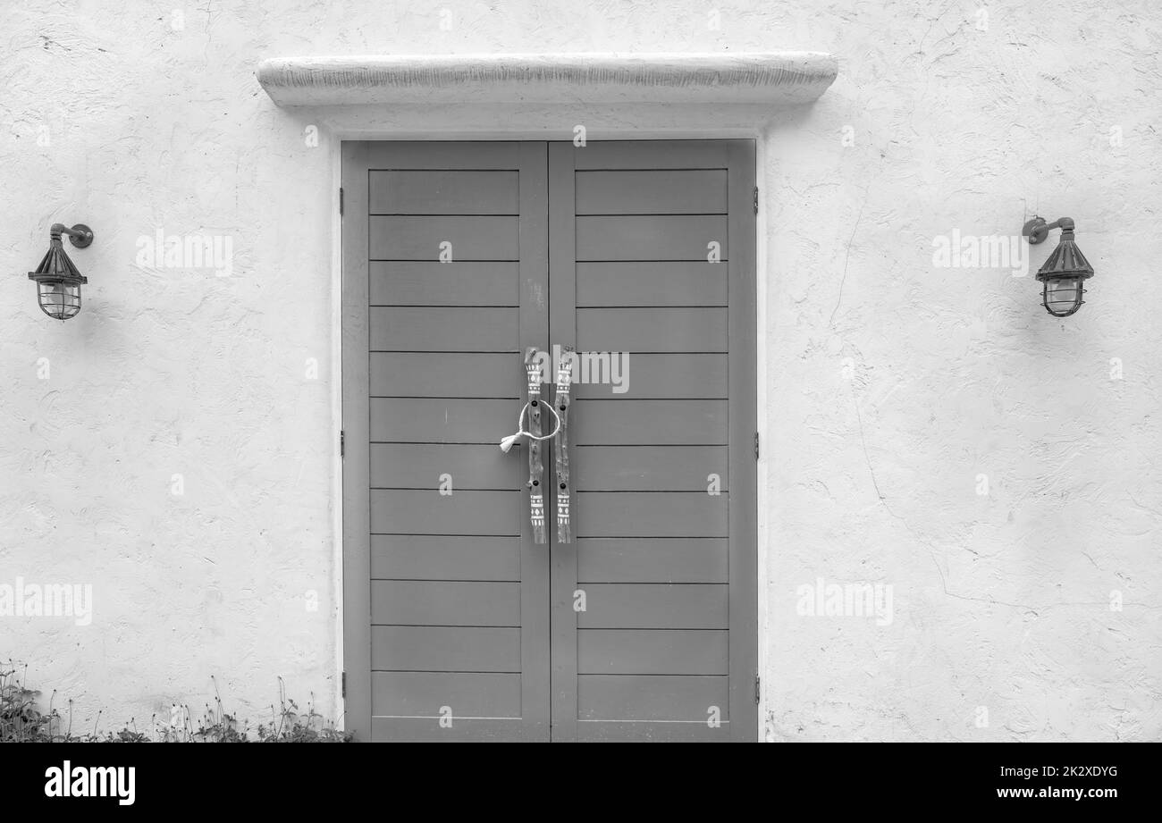 Closed wood door and white concrete wall of the building. Lamp on cement wall near the door. Building exterior. Building wall with rough texture background. Wooden door with handle at the entrance. Stock Photo