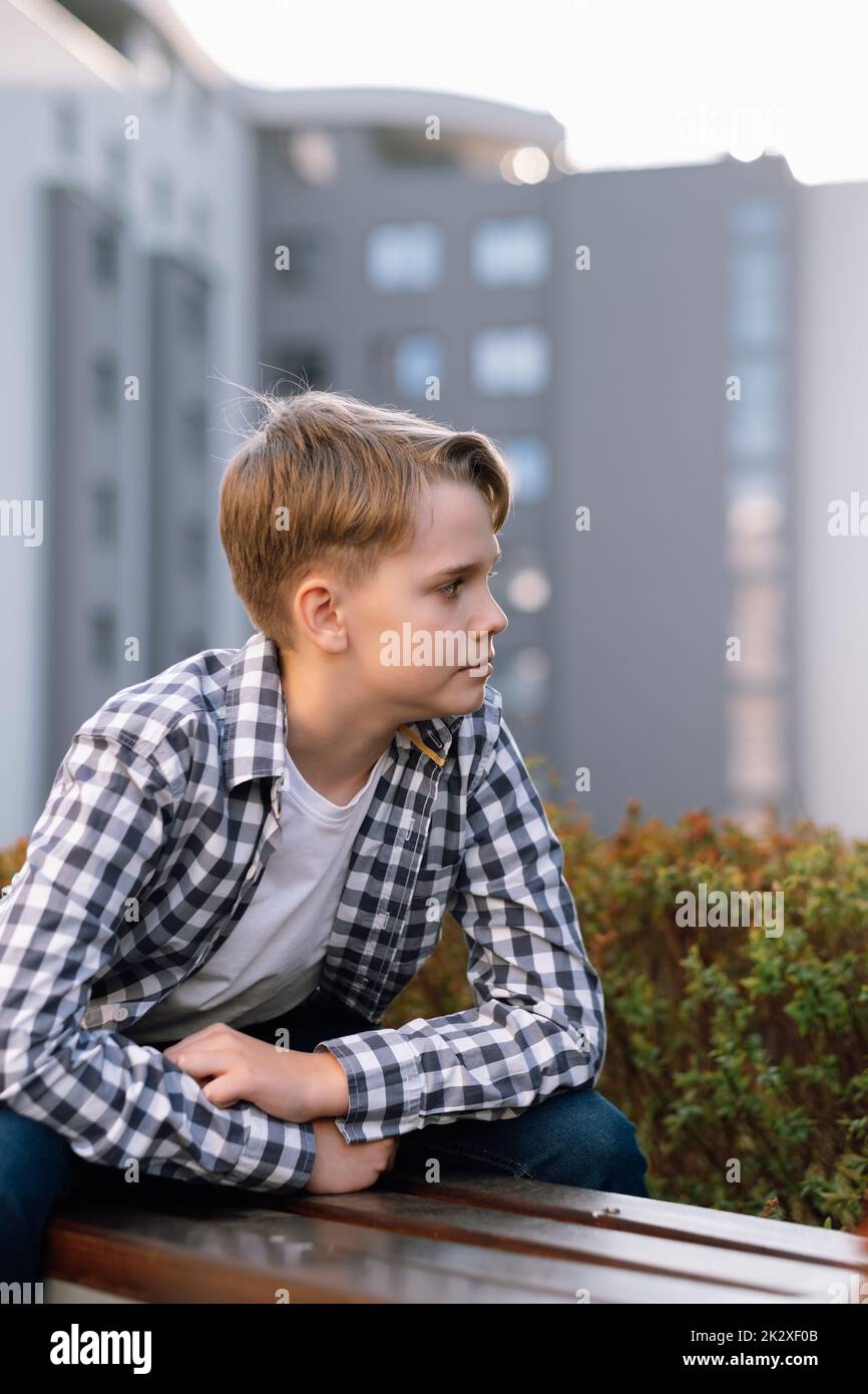 Stylish teenager in casual clothes is relaxing outdoors sitting on a park bench. Stock Photo
