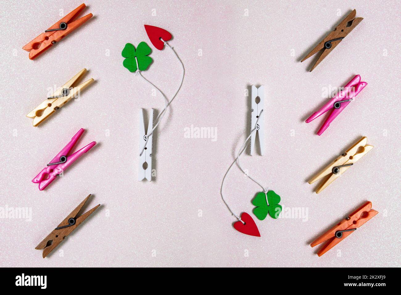 Colorful set of different wooden and plastic clothes pegs on a light pink glitter background. A red and a green lucky clover leaf are attached to each of the white wooden clothes pegs. Top view. Stock Photo