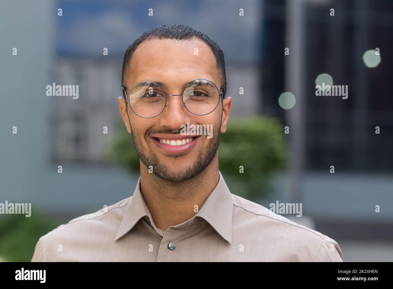 Close-up photo portrait of young entrepreneur wearing glasses, hispanic man smiling and looking at camera, startup entrepreneur outside modern office building. Stock Photo