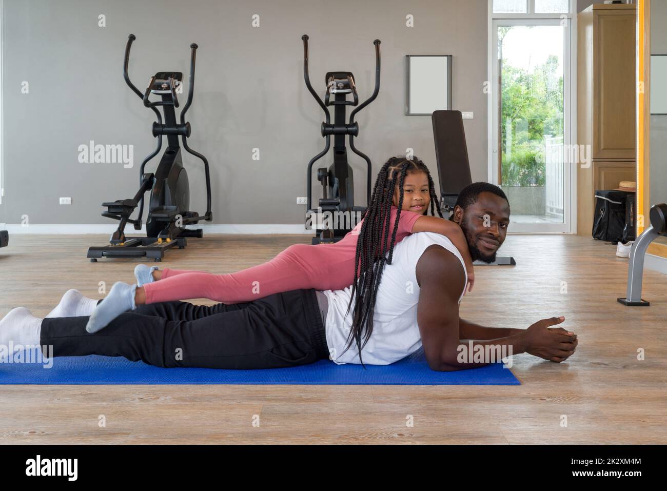 Young short curly black hair man relax on yoga mat while the  daughter are on his back. Happy family enjoy holiday together in fitness center. Stock Photo