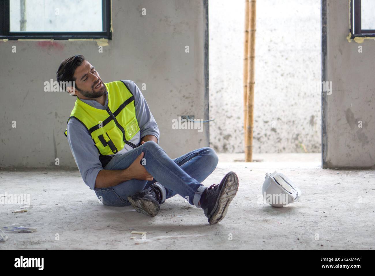 Young caucasian man in safety vest gripped his knee and screams in pain. Due to injuries at the construction site. Stock Photo