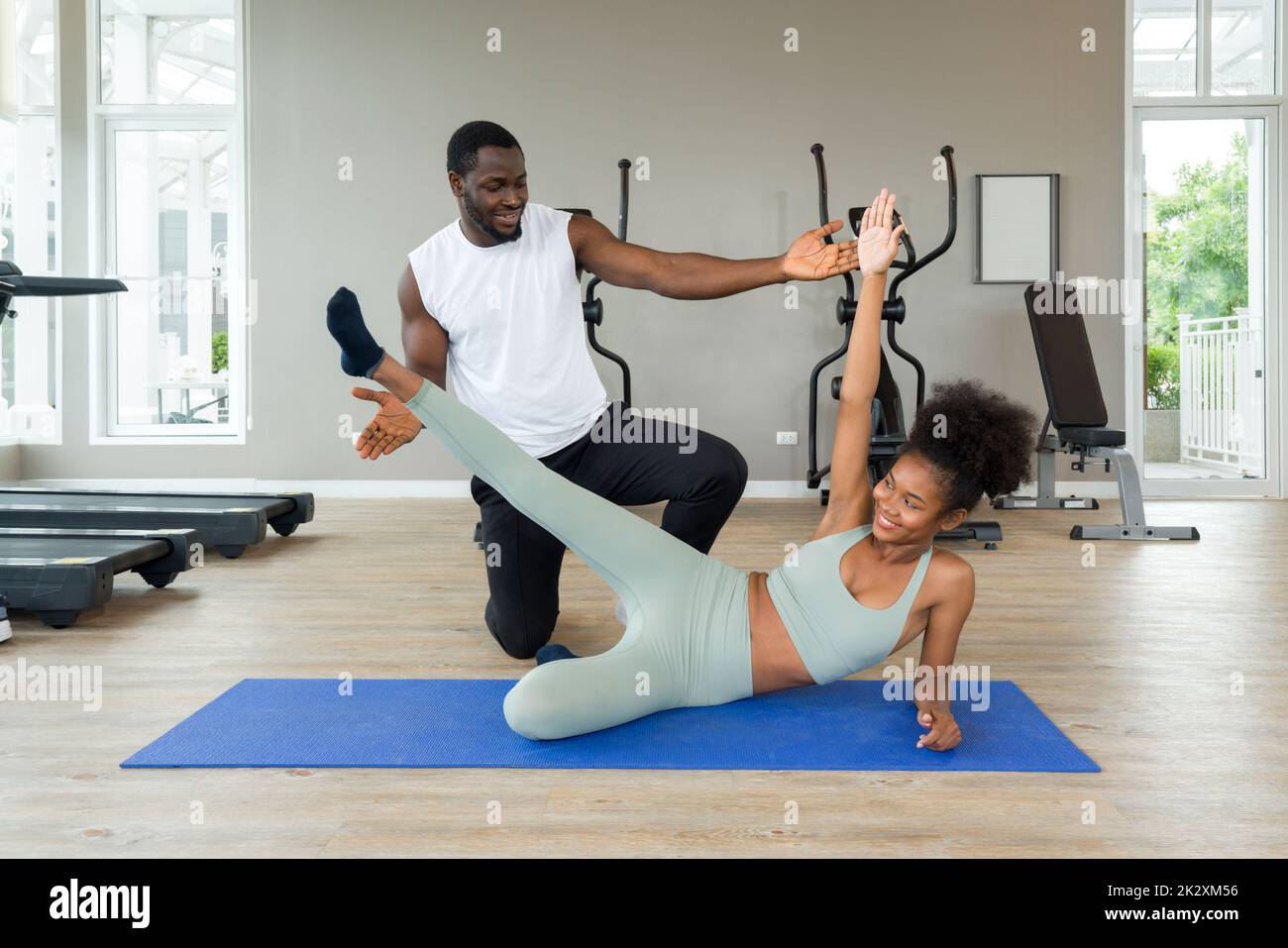 Short curly black hair coach with moustache and beard teach young woman in sportswear how to do side plank crunch. Cardio machines are on the background at the gym. Stock Photo