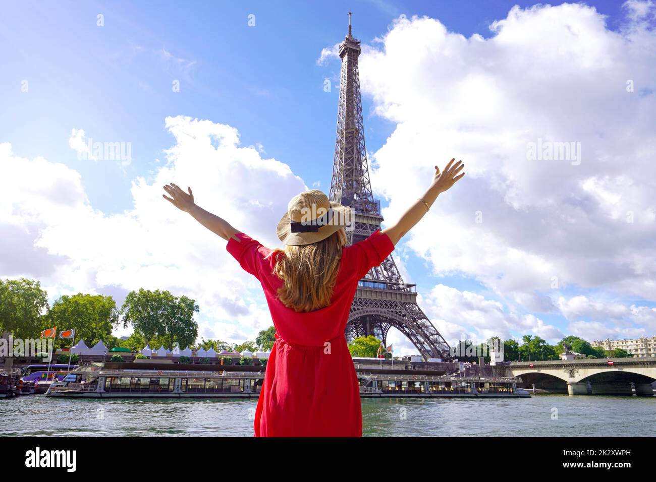 Woman traveler with hat and arms raised on the Seine River in Paris. Back view of fashion woman with open arms in Paris beautiful view with Eiffel Tower on the background. Stock Photo
