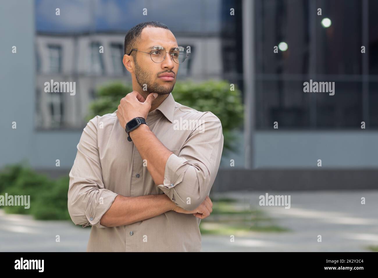 Young pensive businessman in glasses outside modern office building, man thinking and looking away, office worker in shirt. Stock Photo