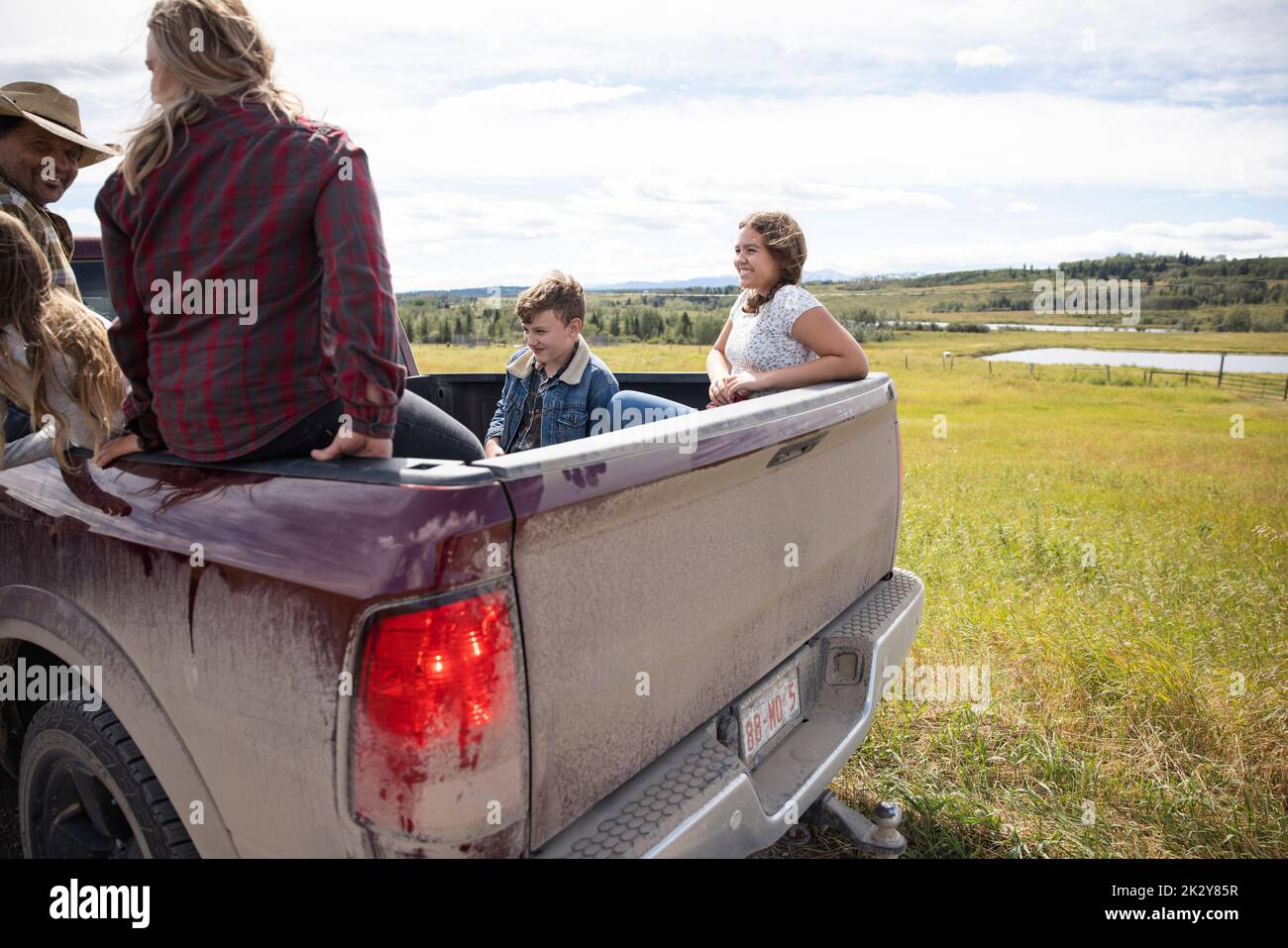 Happy farmer family riding in pickup truck bed on sunny farm Stock