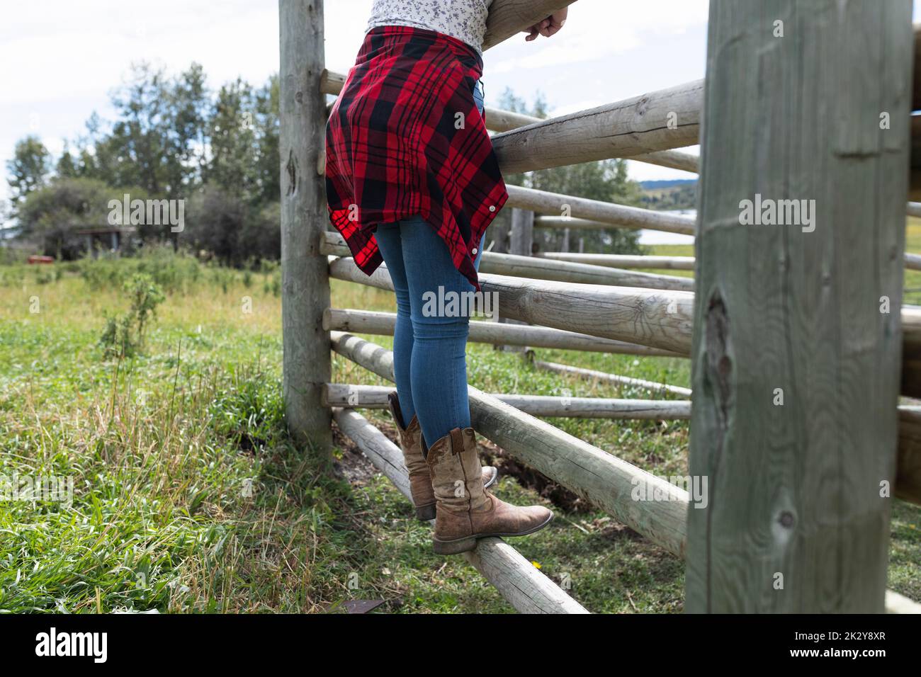 Girl in cowboy boots leaning on wooden fence on sunny rural farm Stock Photo