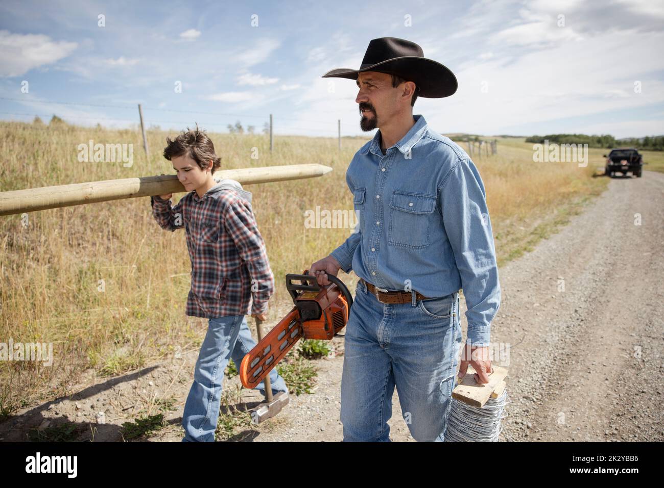 Father and son farmers carrying fence post and chainsaw on farm road