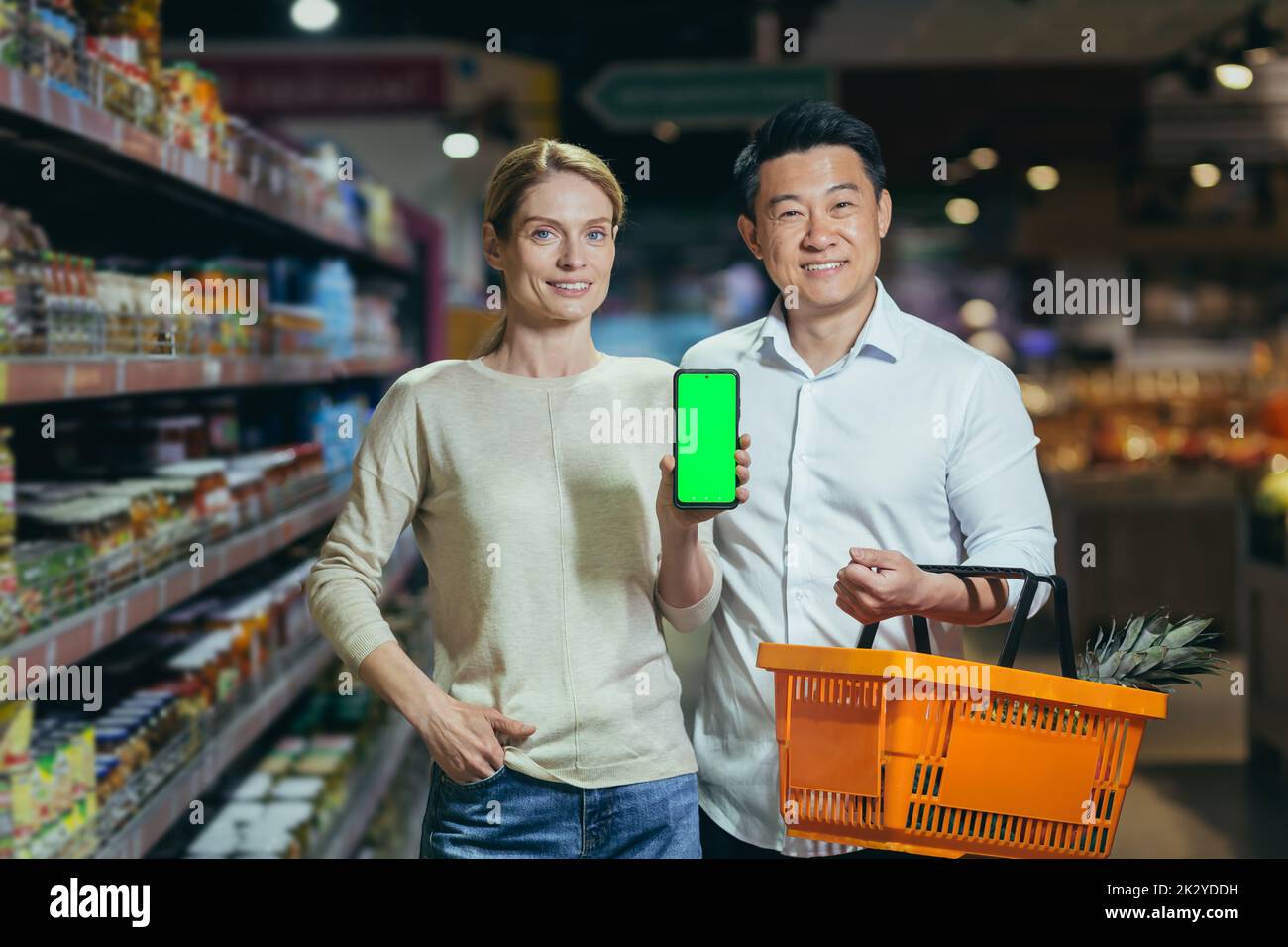 Young family diverse couple of shoppers in supermarket, smiling and looking at camera, grocery department, man and woman holding shopping basket and showing green screen of smartphone. Stock Photo