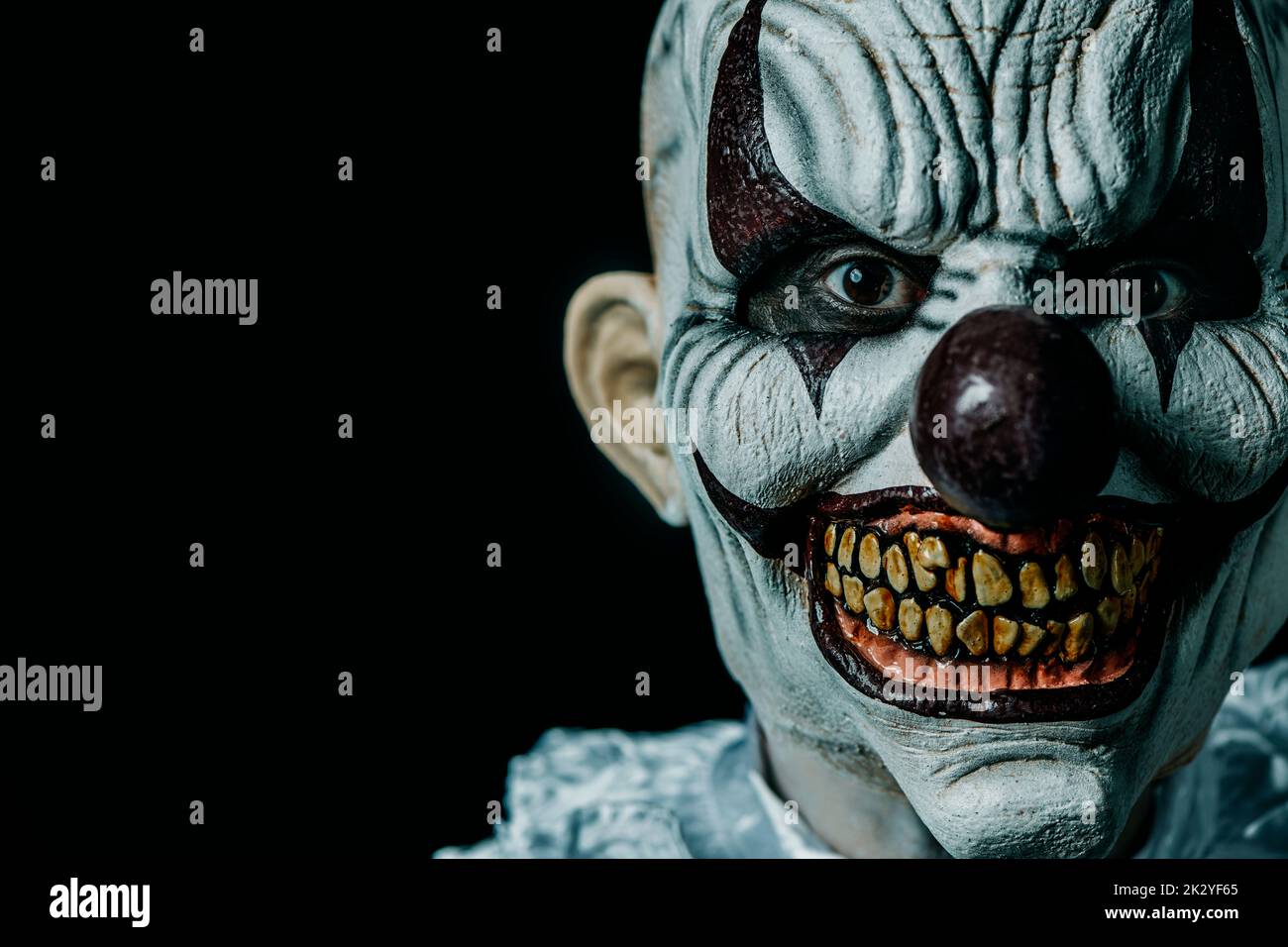 a creepy bald evil clown stares at the observer, against a black background with some blank space on the left Stock Photo
