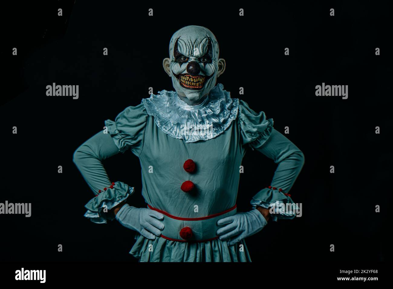 a creepy bald evil clown stands against a black background, with hands on hips, and stares at the observer Stock Photo