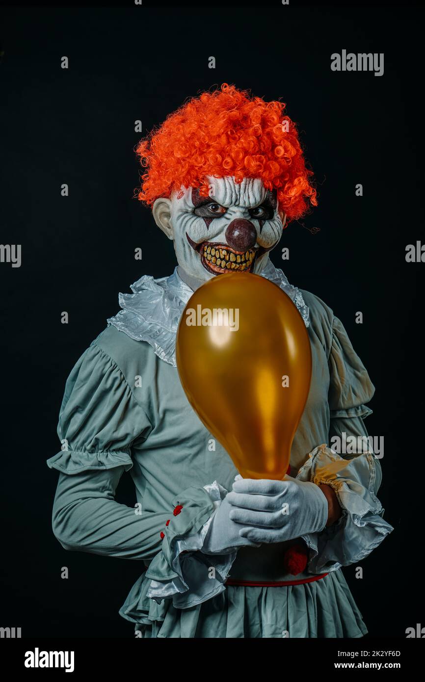 a creepy evil clown with red hair, staring at the observer, holds a golden balloon in front of him, against a black background Stock Photo
