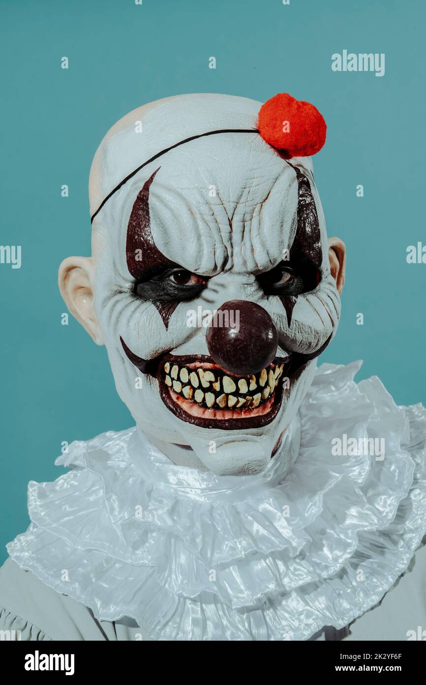 closeup of a creepy bald evil clown, with a menacing smile, wearing a gray costume with a white ruff, and a red pom-pom on his head, on a blue backgro Stock Photo
