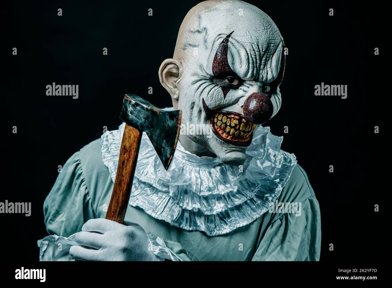 closeup of a creepy bald evil clown threatening the observer with an axe with stains of blood, against a black background Stock Photo