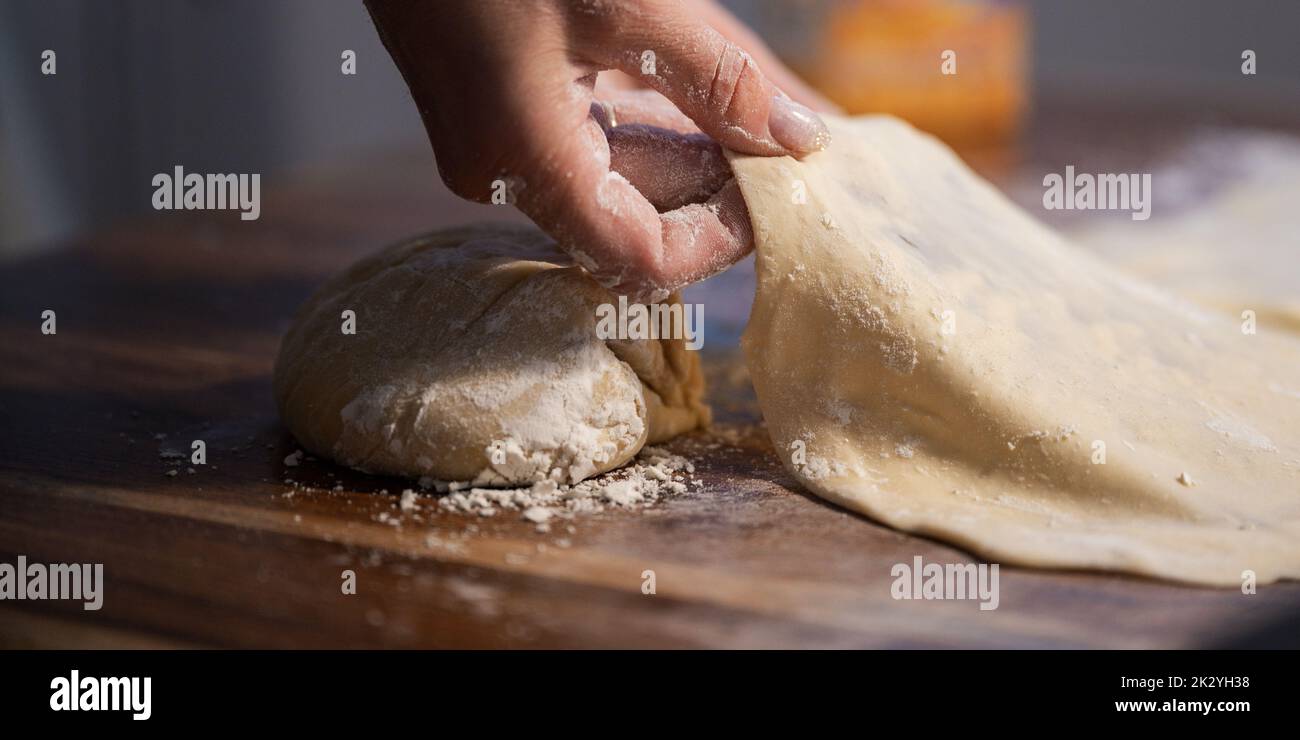 Low angle closeup view of female hands stretching and pulling homemade vegan pastry dough on domestic dining table with another dough bun waiting to b Stock Photo