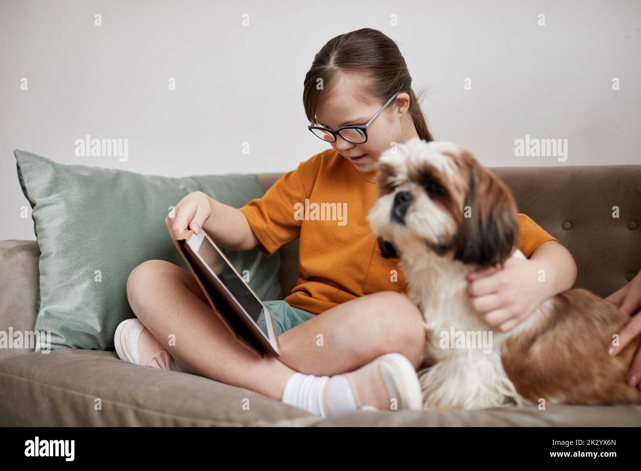 Full length portrait of cute girl with Down syndrome using tablet while sitting on couch with dog companion Stock Photo