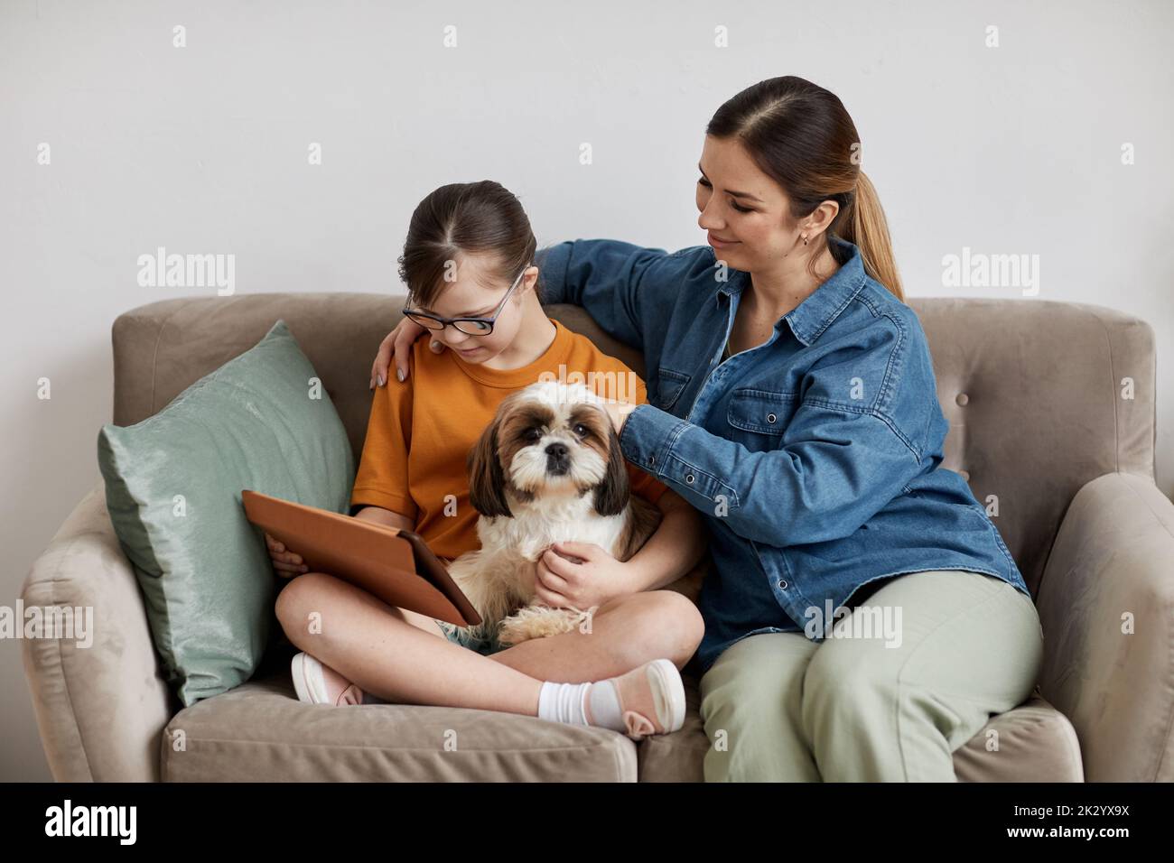 Minimal portrait of caring mother and daughter with Down syndrome sitting on couch with cute dog Stock Photo