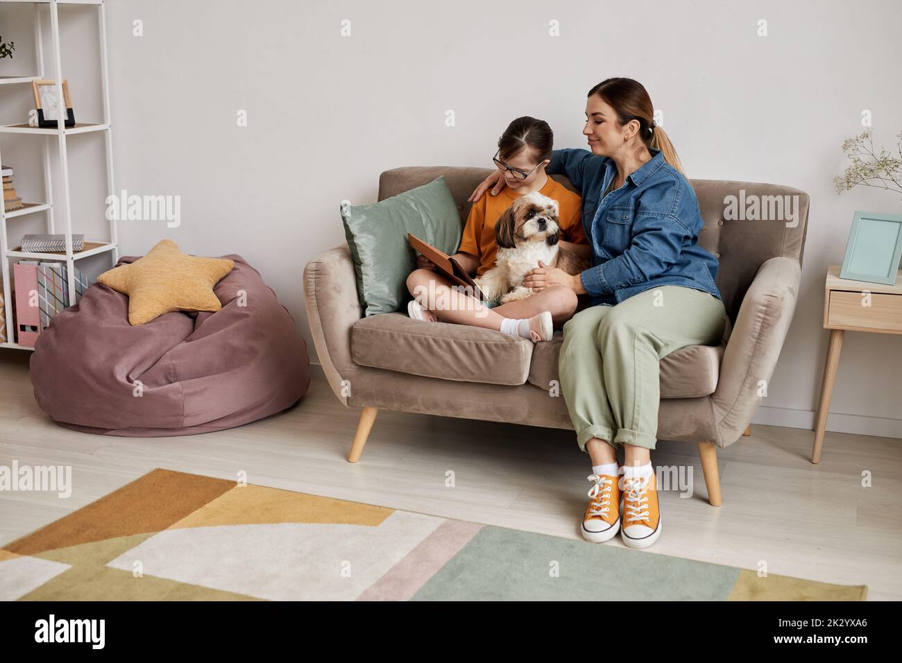 Full length portrait of mother and daughter with Down syndrome using digital tablet together while sitting on couch at home Stock Photo