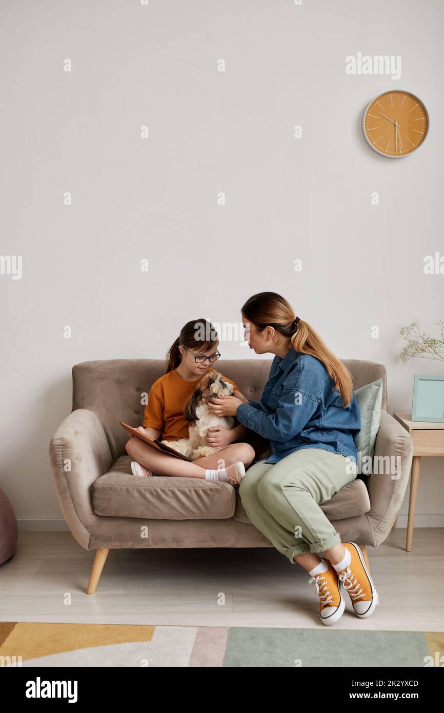 Minimal full length portrait of mother and daughter with Down syndrome playing with dog while sitting on couch at home Stock Photo