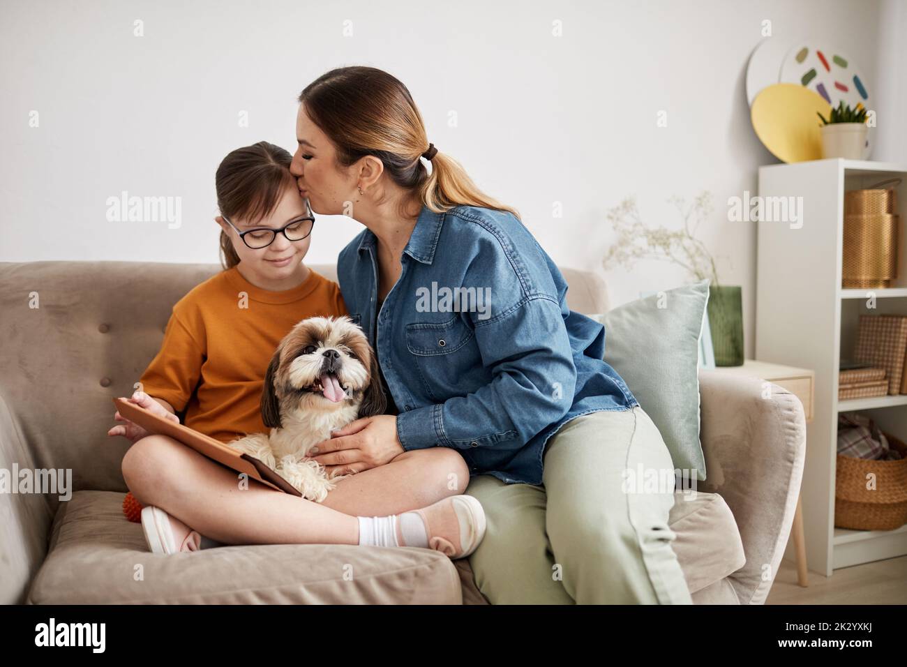 Minimal portrait of loving mother kissing daughter with Down syndrome while sitting on couch at home Stock Photo