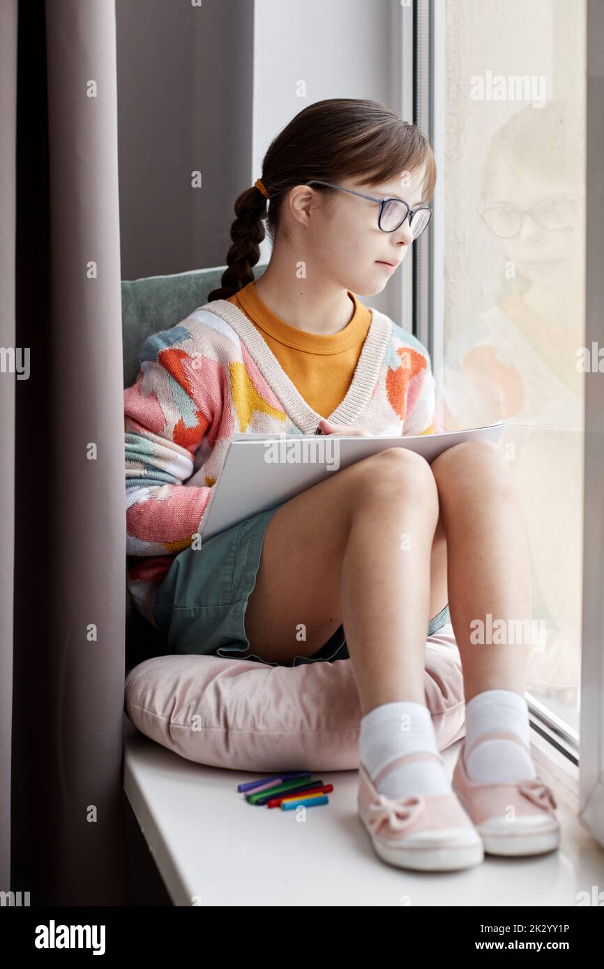 Vertical full length portrait of teenage girl with Down syndrome drawing pictures while sitting by window Stock Photo