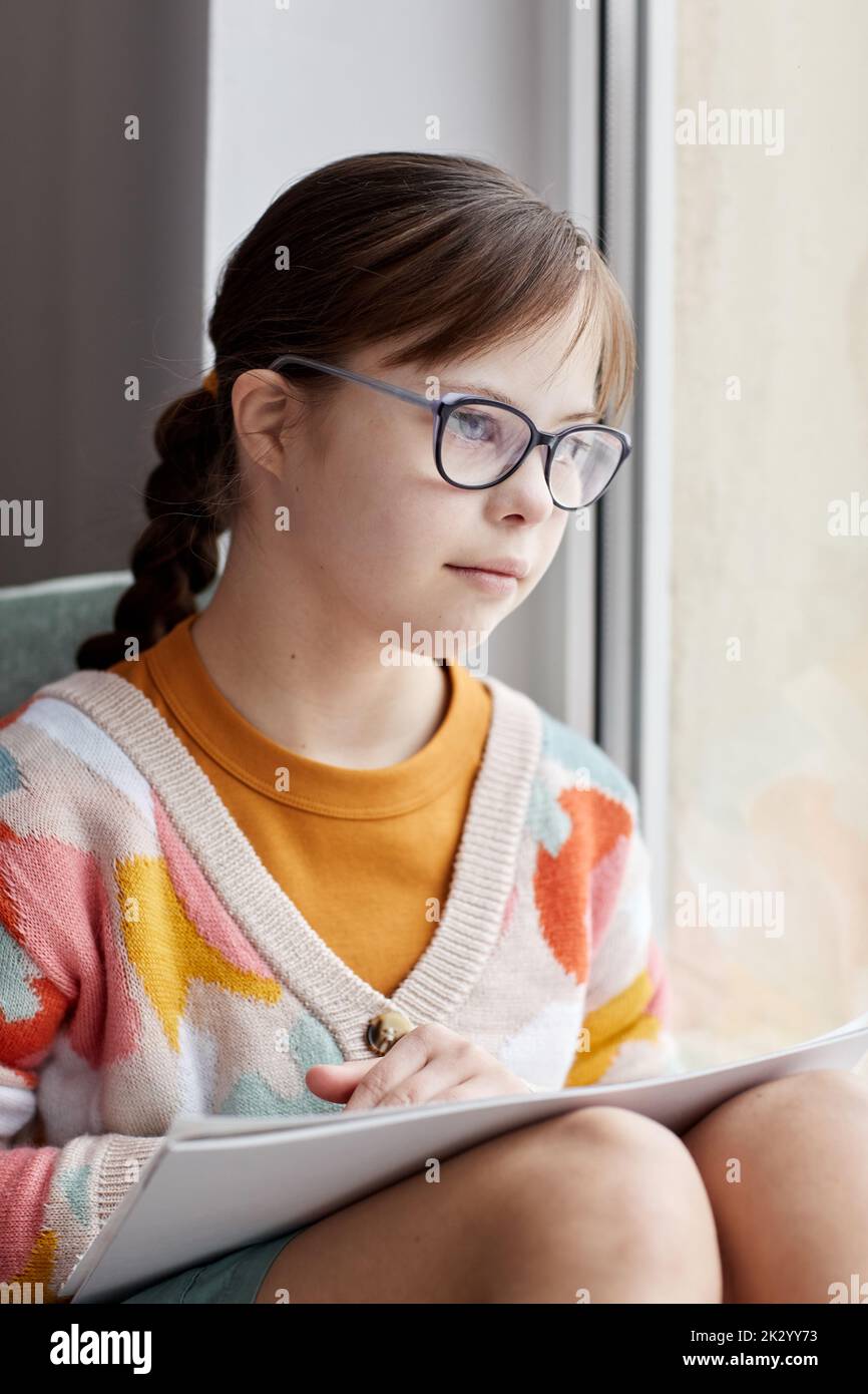 Vertical portrait of teenage girl with Down syndrome drawing pictures while sitting by window at home Stock Photo