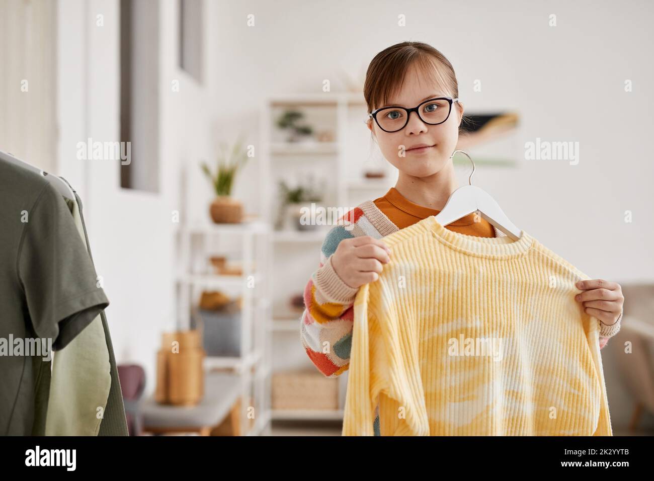 Waist up portrait of teenage girl with Down syndrome choosing clothes and looking at camera, copy space Stock Photo