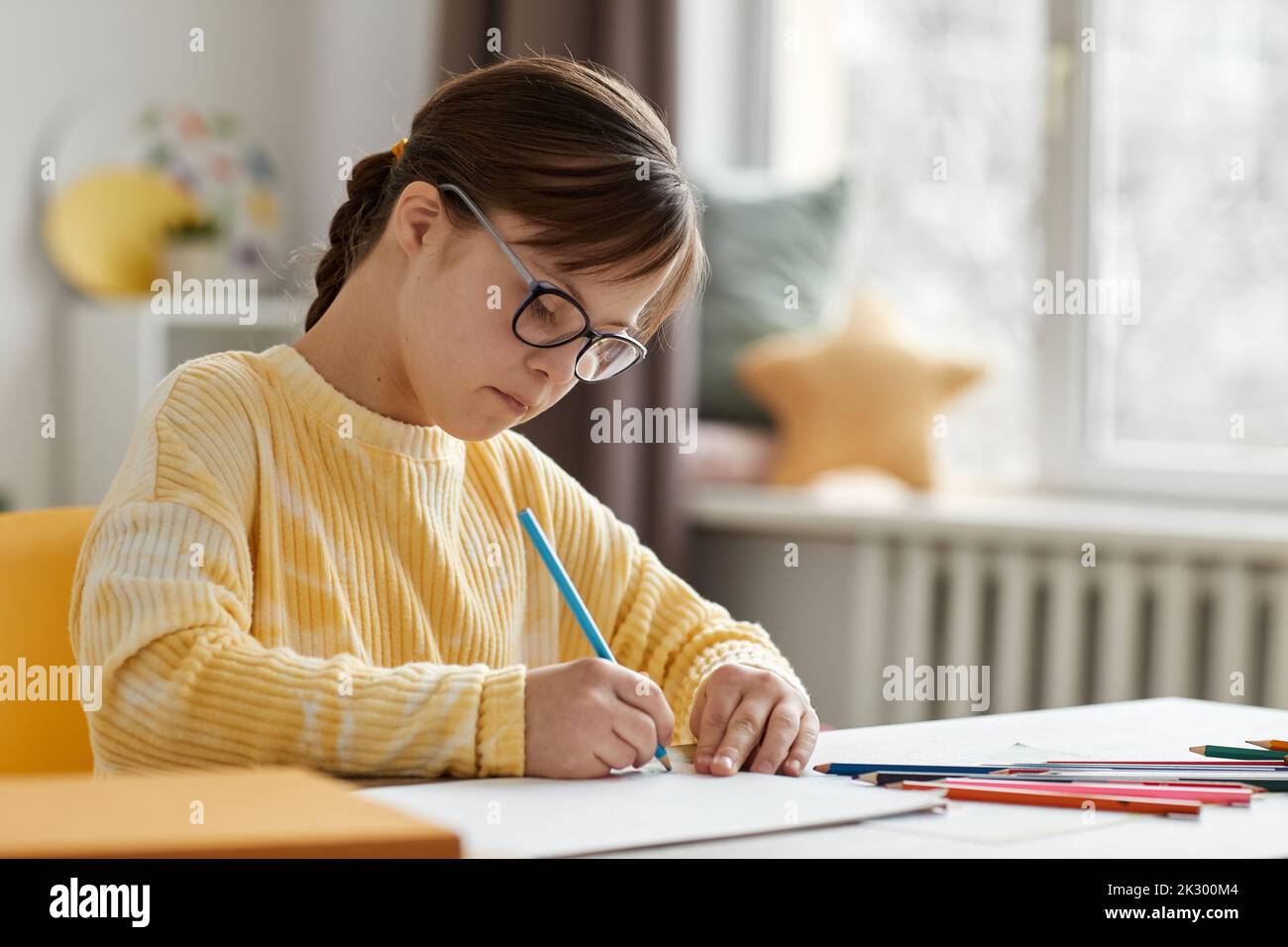 Portrait of cute girl with Down syndrome drawing pictures while sitting at desk at home, copy space Stock Photo