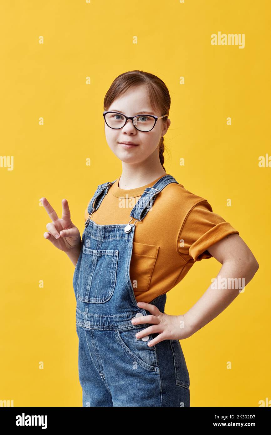 Vertical portrait of teen girl with Down syndrome looking at camera and showing peace sign while standing against yellow background in studio Stock Photo