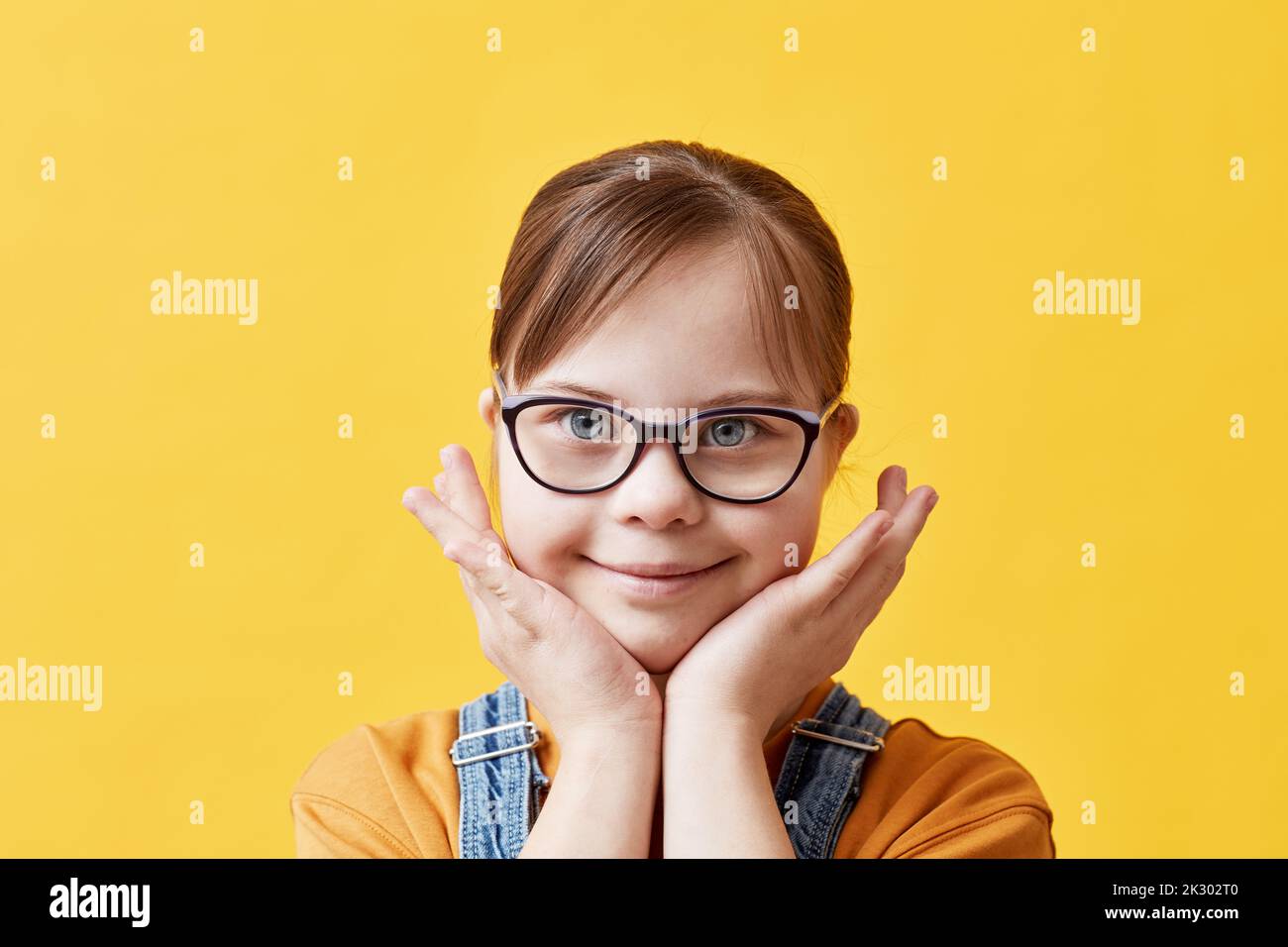 Closeup portrait of cute girl with Down syndrome looking at camera against yellow background in studio Stock Photo