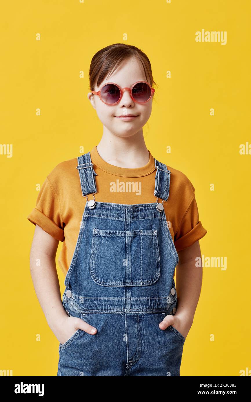 Vertical portrait of cute girl with Down syndrome looking at camera against yellow background and wearing sunglasses Stock Photo