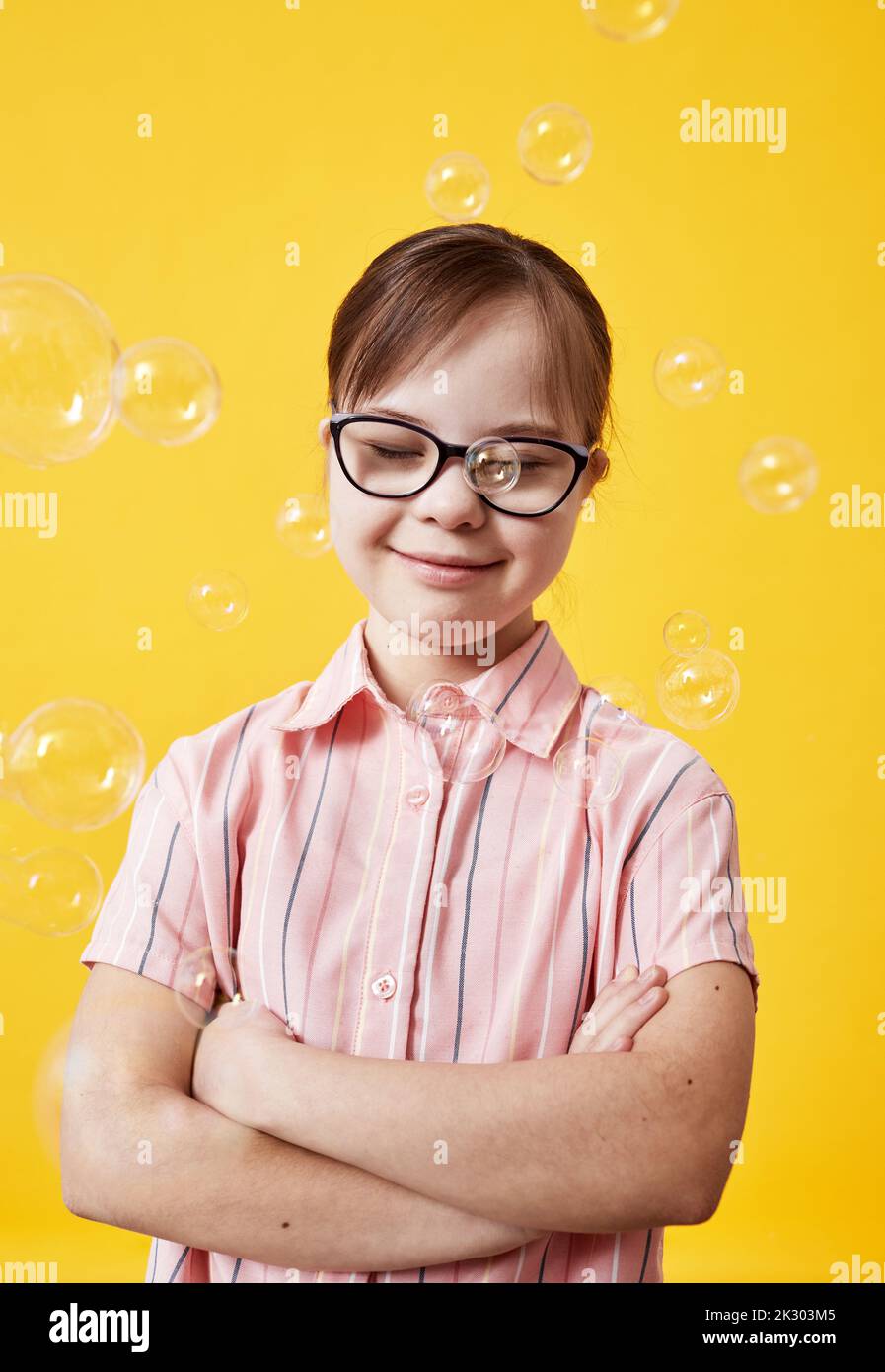 Vertical portrait pf playful cute girl with Down syndrome playing with bubbles against yellow background Stock Photo