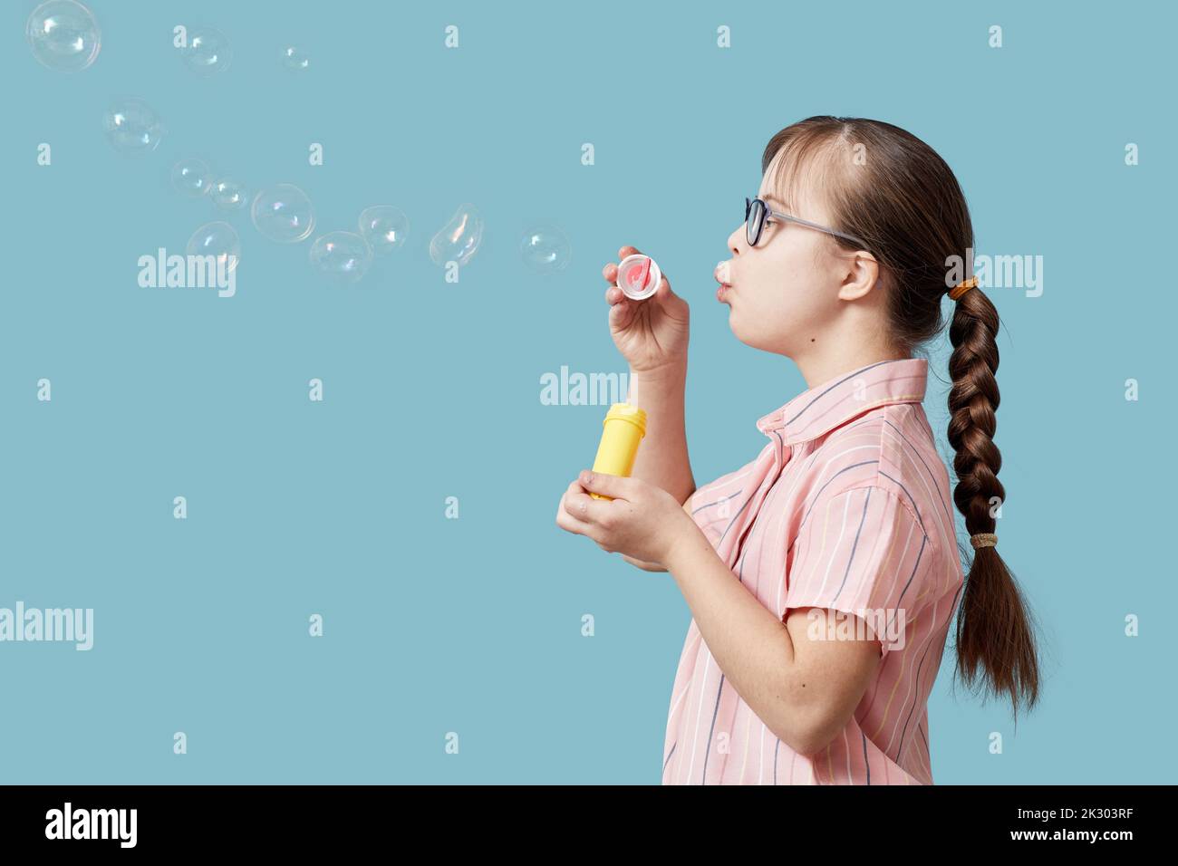 Side view portrait pf playful girl with Down syndrome blowing bubbles against blue background Stock Photo