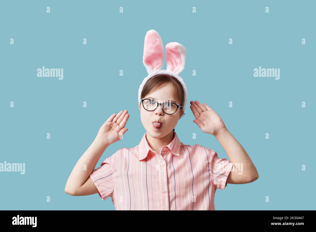 Portrait of teen girl with Down syndrome wearing Easter bunny ears against blue background and making funny faces Stock Photo