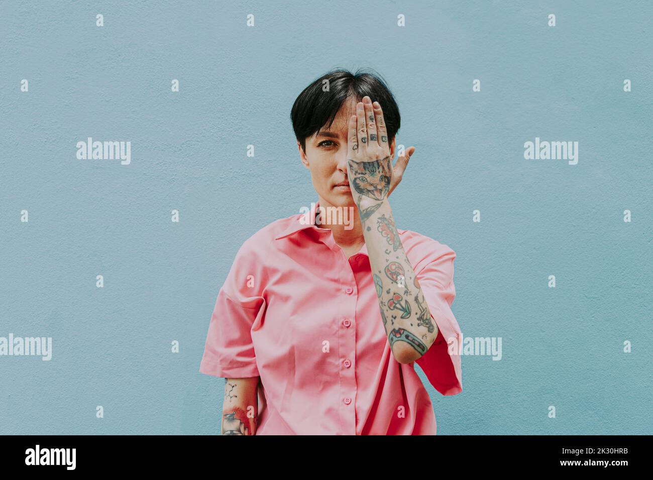 Tattooed woman covering eyes with hand standing in front of blue wall Stock Photo