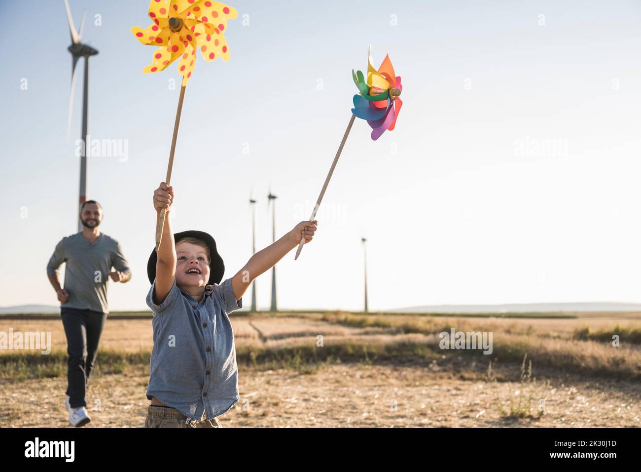 Boy with colorful pinwheels running in windpark with father following Stock Photo