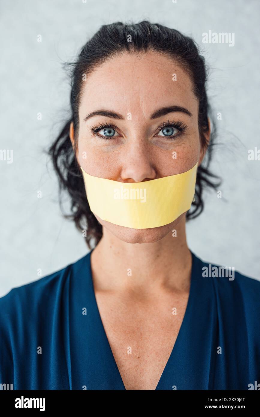 Woman with tape over mouth standing in front of gray wall Stock Photo