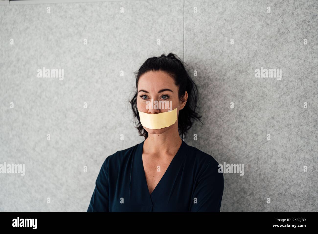 Woman with tape over mouth standing in front of gray wall Stock Photo