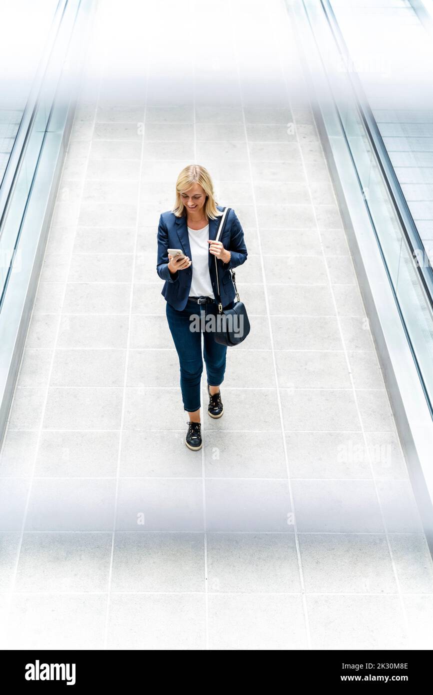 Smiling businesswoman using phone walking on walkway Stock Photo