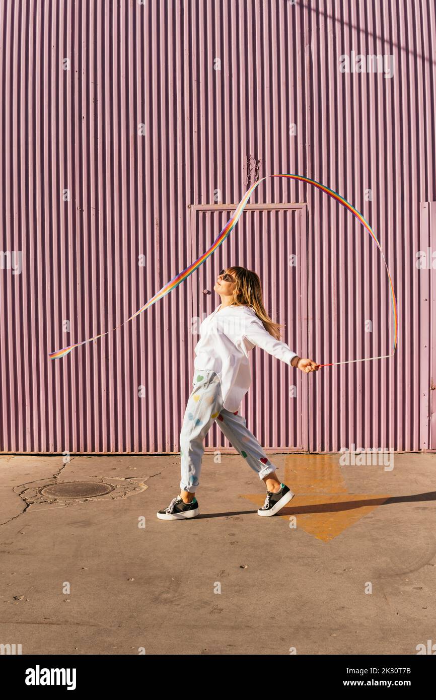 Carefree woman doing rhythmic gymnastics with rainbow colored ribbon in front of corrugated wall Stock Photo